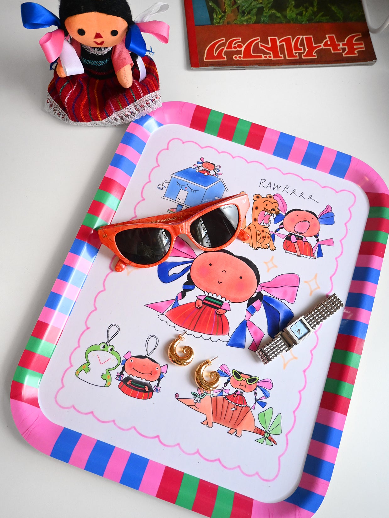 Colorful tray with cartoon designs, sunglasses, and a watch on a white surface. Design by Natali Koromoto.