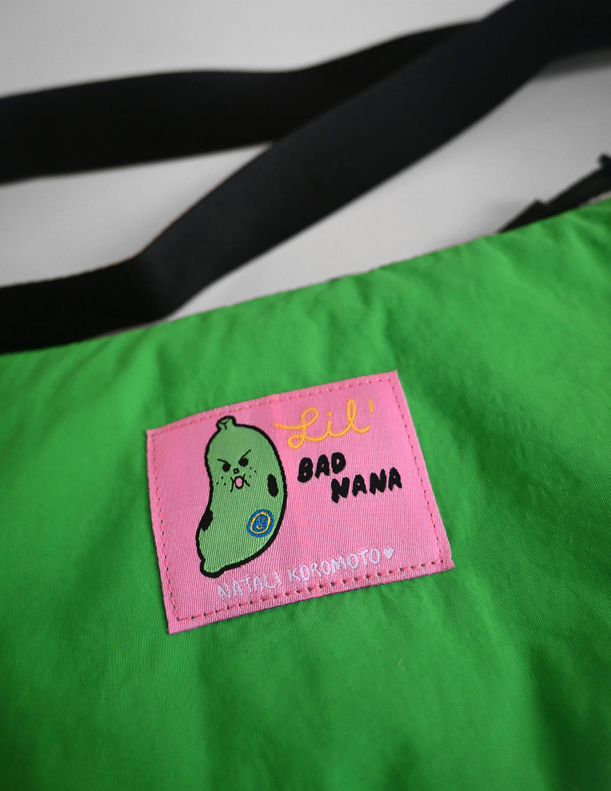 "Bad-nana" Crescent Bag. Natali Koromoto design NYC.
- A colorful cartoon illustration of a green banana-like character with an angry face and the words 'Lil' BAD NANA' on a solid pink background.
