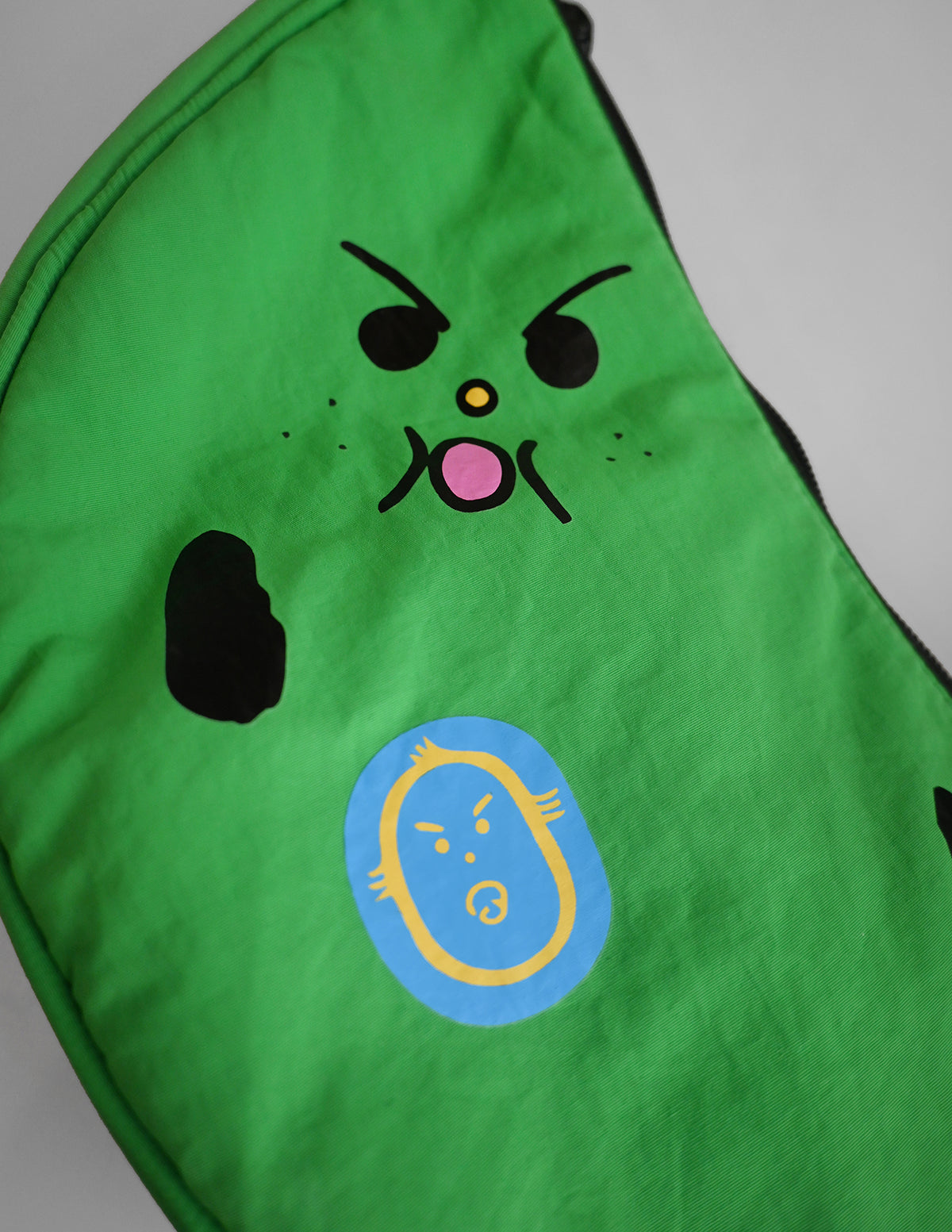 "Bad-nana" Crescent Bag. Natali Koromoto design NYC.
- A colorful cartoon illustration of a green banana-like character with an angry face and the words 'Lil' BAD NANA' on a solid pink background.