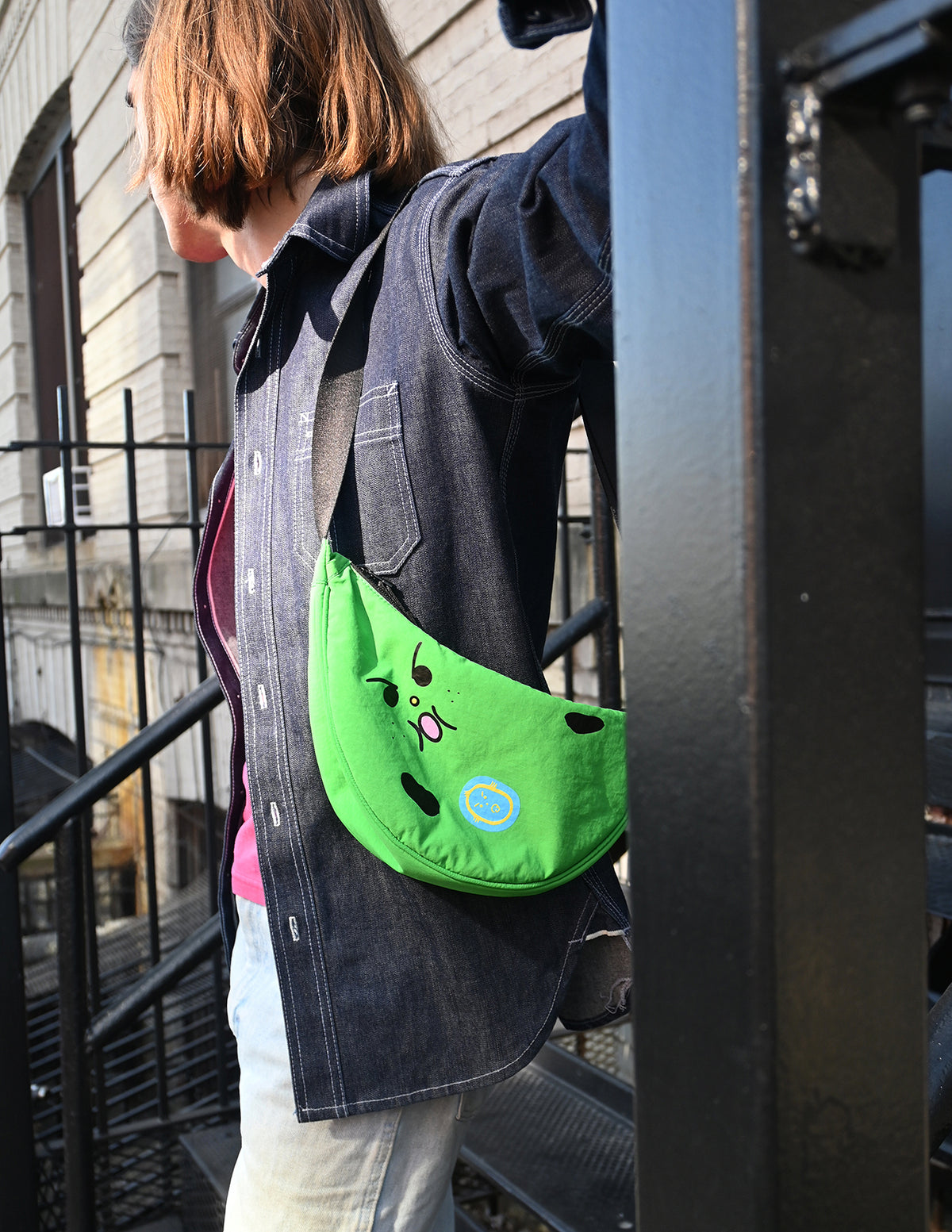 "Bad-nana" Crescent Bag. Natali Koromoto design NYC.
- A colorful cartoon illustration of a green banana-like character with an angry face and the words 'Lil' BAD NANA' on a solid pink background.