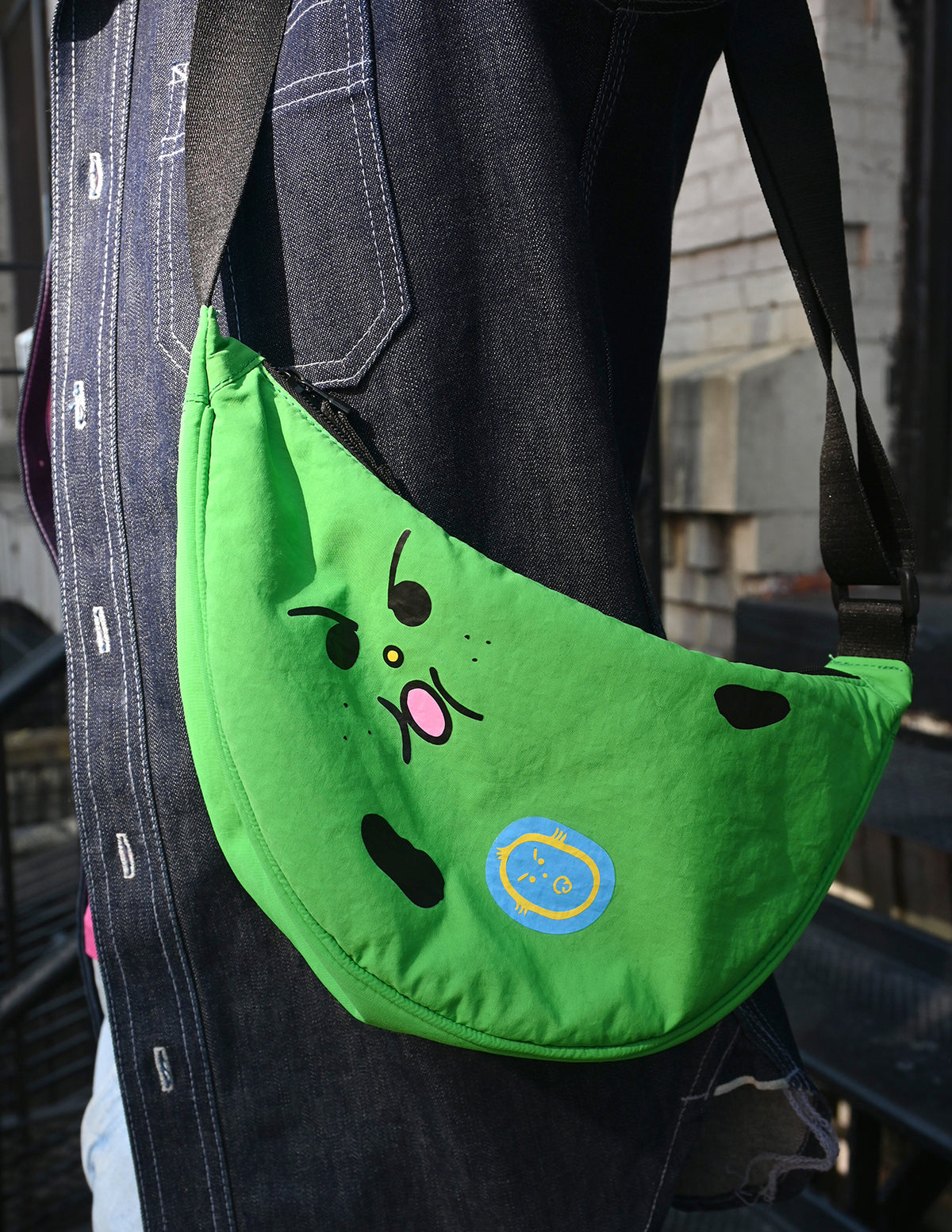 "Bad-nana" Crescent Bag. Natali Koromoto design NYC.
- A colorful cartoon illustration of a green banana-like character with an angry face and the words 'Lil' BAD NANA' on a solid pink background.